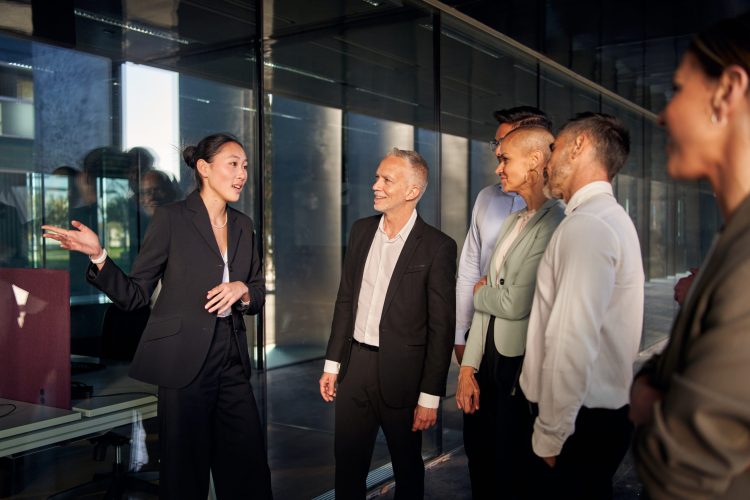 Team meeting diverse professionals listening to Chinease female team leader. Multiracial smiling colleagues gathered inside hallway business building. Group office workers attending Asian woman boss.